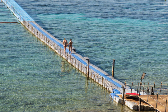 High angle view of two playful young female friends in bikinis walking and flirting on a long floating pontoon bridge in the shallow clear Red Sea water at a luxury resort in Sharm El Sheikh, Egypt