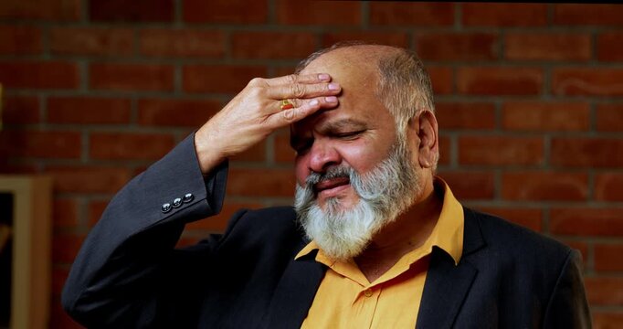 Indian senior businessman or corporate executive suffering from a painful headache and touching his temples while standing against a red brick wall in a cozy office with moody lighting.