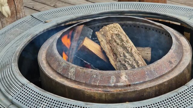 Wooden logs burn in a modern metal fire pit (pit ring) on ​​a wooden terrace. Setting up a fire pit on the street, in the garden or at the cottage. Fire in a metal fire pit outdoors. Coziness and rela
