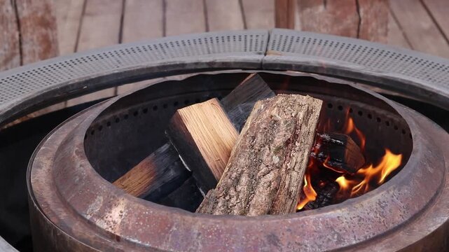 Wooden logs burn in a modern metal fire pit (pit ring) on ​​a wooden terrace. Setting up a fire pit on the street, in the garden or at the cottage. Fire in a metal fire pit outdoors. Coziness and rela