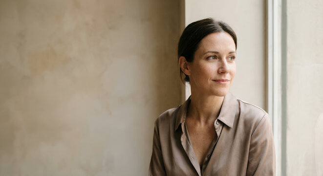 A woman in a tan silk blouse looks out a window.
