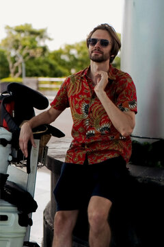 Stylish man in batik shirt and sunglasses sitting outdoors at airport