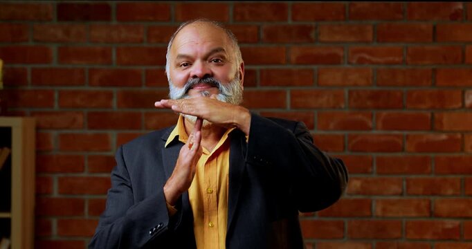 Indian senior businessman or corporate executive gesturing a time out with his hands signaling for a break or a stop in proceedings while standing against a red brick wall in a cozy moody office