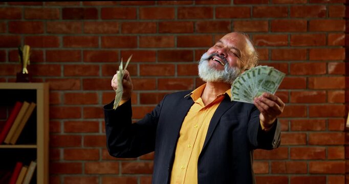 Indian senior businessman flaunting a fan of five hundred rupee notes to the camera with joy and success while standing against a red brick wall in a cozy office with professional moody lighting