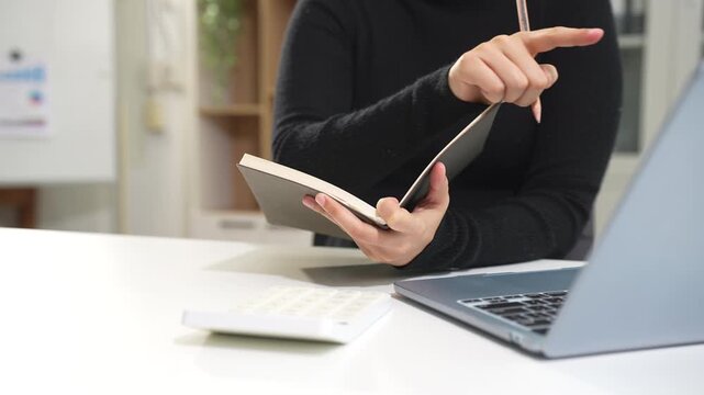 Employee, taking notes sitting at desk using  laptop searching for information at work.