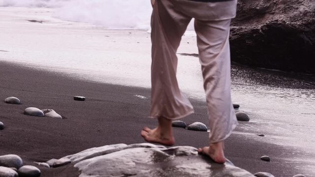 Extreme close-up horizontal shot of a woman's bare feet walking on wet black volcanic sand and rocks with white sea foam in the background
