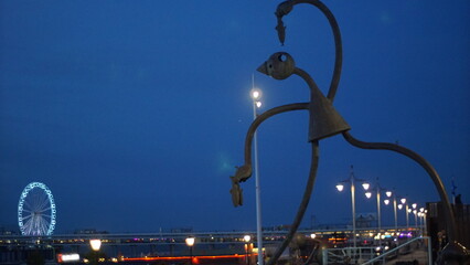 Tall abstract metal sculpture stands beside a riverside promenade at dusk, illuminated by streetlights, with a glowing Ferris wheel and bridge in the background. © Aperezer