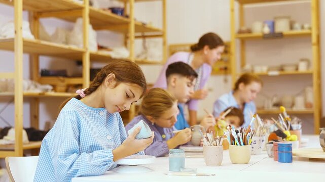 Teenagers and children at a ceramics workshop in an art studio. Woman teacher shows how to paint clay dishes with a brush