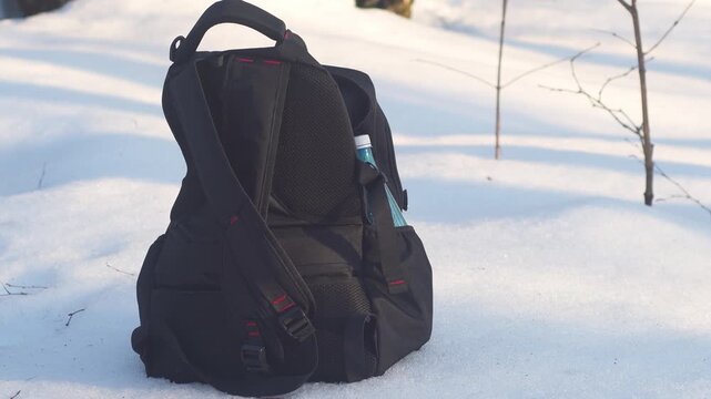 A simple black backpack stands in the snow. An empty plastic bottle is in the backpack pocket. Hiking in winter weather.