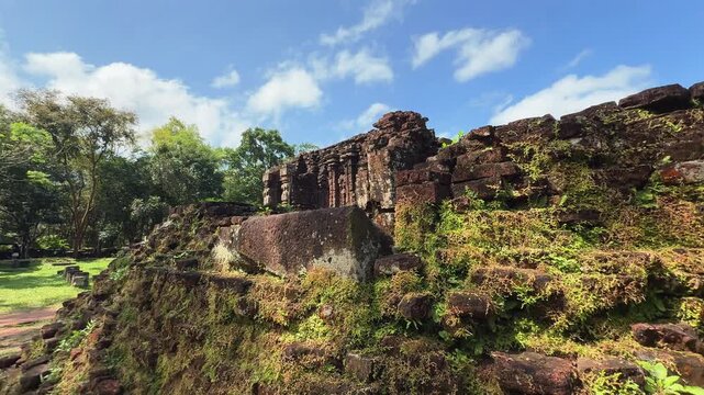Dramatic tilt-up reveal of ancient Cham temple ruins at My Son Sanctuary near Hoi An, Vietnam. Moss-covered sandstone towers rise against blue sky amid lush tropical jungle.