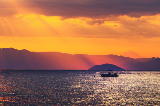 Silhouette of a boat on calm sea during vibrant sunset, with layered mountain ranges in the background and dramatic sun rays through the clouds.