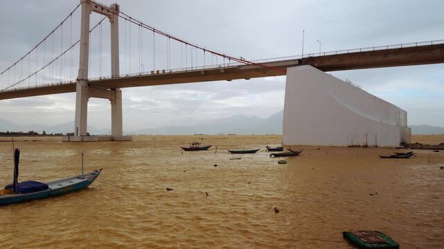 Suspension Bridge towering above Brown River with Fishing Boats anchored near Shore in Wide Angle under Cloudy Mountains creating Serene Coastal Mood, Riverside Travel Concept