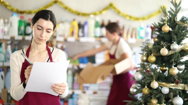 Attentive saleswoman recording stocks on paper when other employee arranges racks in supermarket during Christmas 