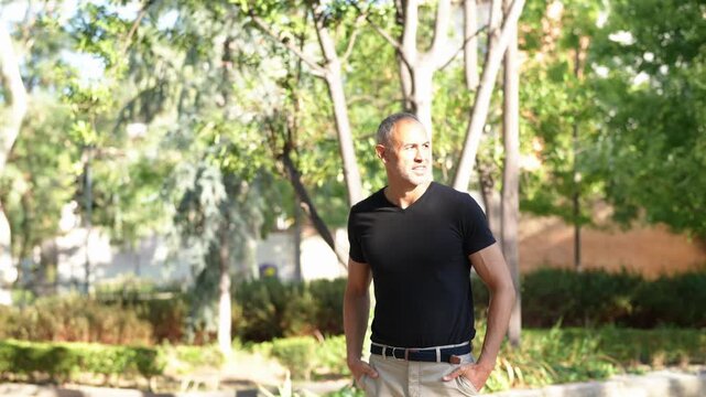 Man stands calmly in a sunlit garden. He looks relaxed, with hands on hips. Trees sway gently behind him. Soft daylight creates warm, inviting atmosphere. Perfect for lifestyle, travel