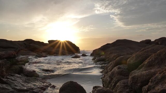 Amber sunbeams during sunrise pierce through bruised clouds over the rocky coastline as ocean waves come rolling into an inlet towards the camera.