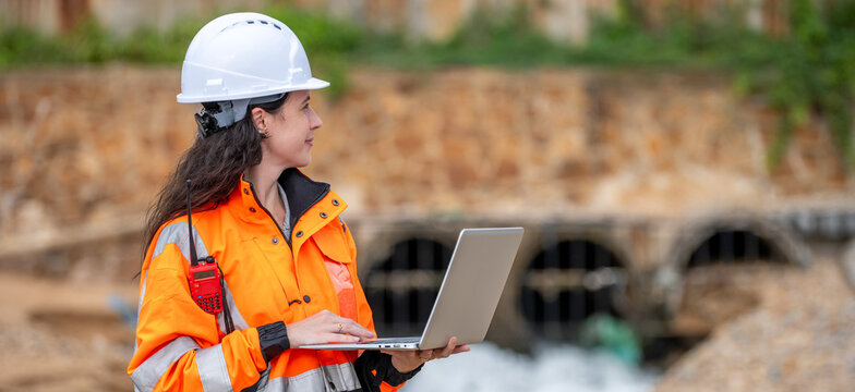 Female environmental scientist in safety gear standing and using laptop and radio for monitoring ecological impact inspection near industrial gas storage tank at fuel terminal site.