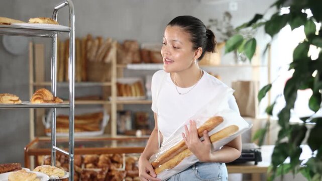  Asian woman looks at rack shelf showcase filled with various flour products, bread assortment, croissants, puffs with filling, choosing something in bakery. Quick snack on go