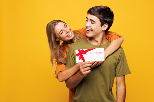 Young fun couple two friends family man woman they wears casual clothes together hold gift certificate coupon voucher card for store hug isolated on plain yellow background studio. Lifestyle concept