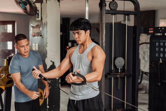 Young Asian man does low cable crossover at the gym, guided by a trainer to improve chest activation, form, and controlled resistance for muscle development.