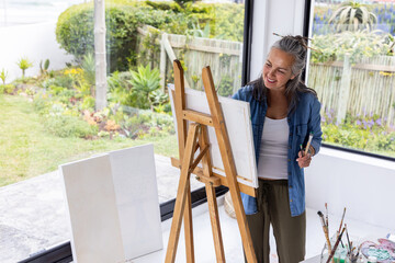 Naklejka premium Senior woman wearing denim shirt, olive pants standing at easel painting with palette, copy space