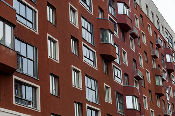 Fototapeta premium Red brick apartment building rises tall. Windows reflect sky and clouds. Balconies curve around each unit. Building shows urban residential life. Ideal for cityscape or housing themes