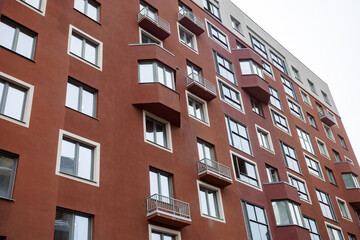 Fototapeta premium Red brick building with many windows and balconies. Balconies feature metal railings, some closed, others open. Windows reflect sky and clouds, adding depth. Architecture shows urban living