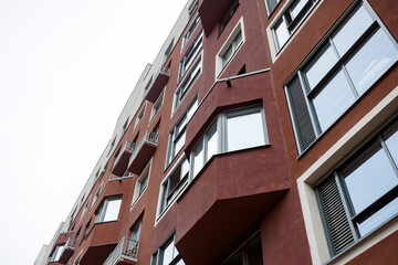 Fototapeta premium Red brick building rises against pale sky. Windows reflect clouds and daylight softly. Balconies offer quiet urban charm. Ideal for architecture, cityscape, or modern living themes