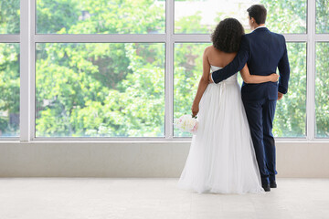 Obraz premium Romantic newlywed couple embracing and looking through large window, bride and groom from behind in bright modern interior with green garden view and copy space