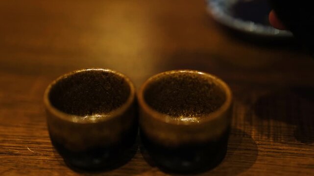 Pouring sake into small traditional Japanese cups on wooden table, cinematic slow motion.
