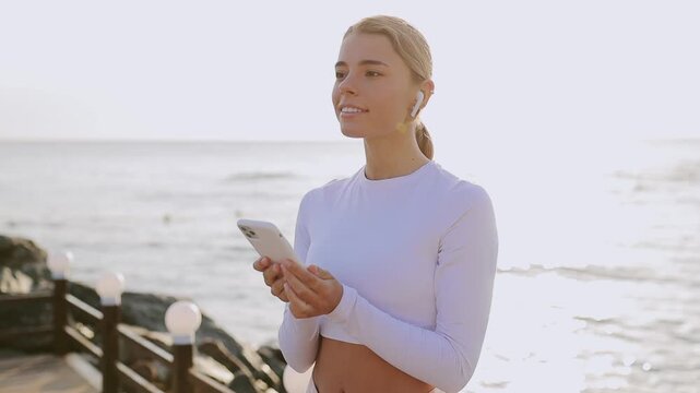 Young strong sporty fit sportsman woman wear sports clothes listen to music in earphones use mobile cell phone warm up train at sunrise sun over sea beach outdoor on pier seaside in summer day morning