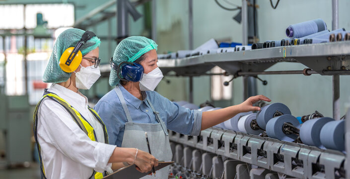 Textile inspection factory teamwork female supervisor in safety vest checking and discussing yarn production quality with woman worker in textile mill holding thread spool and clipboard.