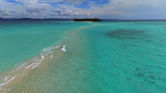 Aerial view shows a tropical island with clear turquoise waters. Narrow sandbar connects the island to the mainland. White waves gently break near the shore. Sky has scattered clouds with blue patches