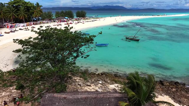 Top-down view of paradise beach. Palm fronds frame crystal clear water. Wooden boats float near sandy shore. Visitors sunbathe or paddle in calm ocean. Perfect for tourism, advertising