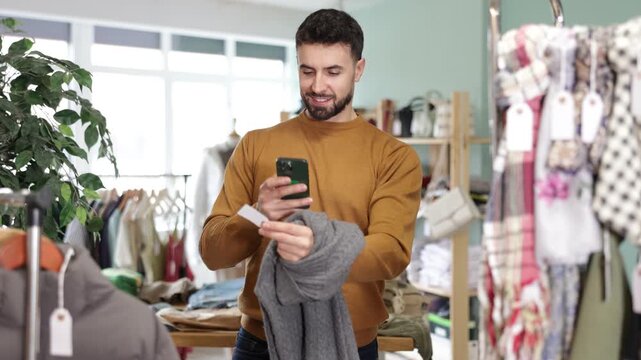 Young man buyer scanning qr code for warm sweater in clothing store