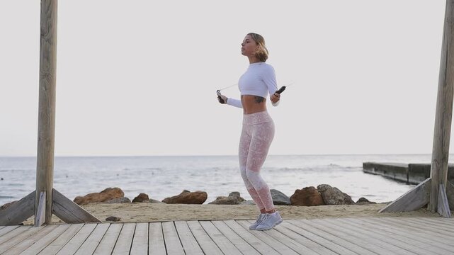 Full body young strong sporty athletic fit sportsman woman wear sports clothes jumping on skipping rope warm up train at sunrise sun dawn over sea beach outdoor on pier seaside in summer day morning