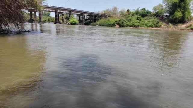 Serene river flows beneath bridge, with calm waters reflecting lush greenery and trees, creating tranquil outdoor landscape that embodies beauty of nature.