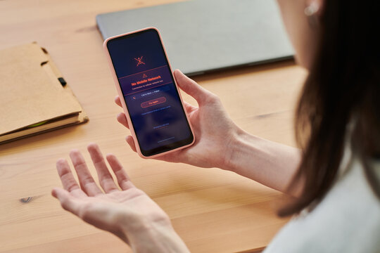 Young adult woman holding smartphone showing no mobile network error while working at desk, expressing frustration. Ideal for telecom outage, connectivity problem, remote work disruption themes