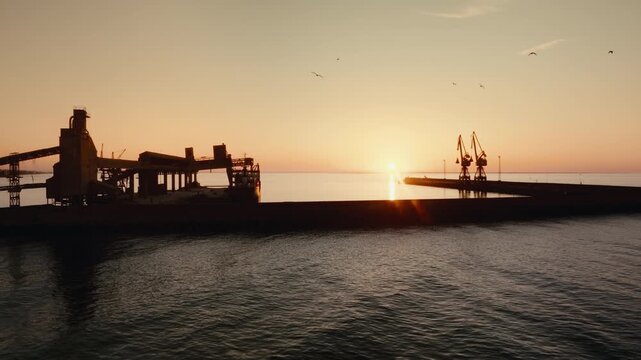 Epic 4K drone aerial pulling back over water and industrial harbor pier at sunset. Cargo cranes and calm water Ideal for shipping, logistics, maritime business, trade, and cinematic industrial scenes.