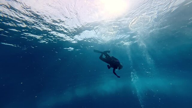 Solitary scuba diver descends through crystal clear sapphire depths towards a mysterious ocean floor as bright midday sunbeams pierce the shimmering surface in this wide low angle summer adventure