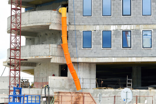 Building under construction, orange debris chute for waste disposal against grey wall facade. Construction site, waste management,  plastic debris tube attached to building balcony, garbage removal