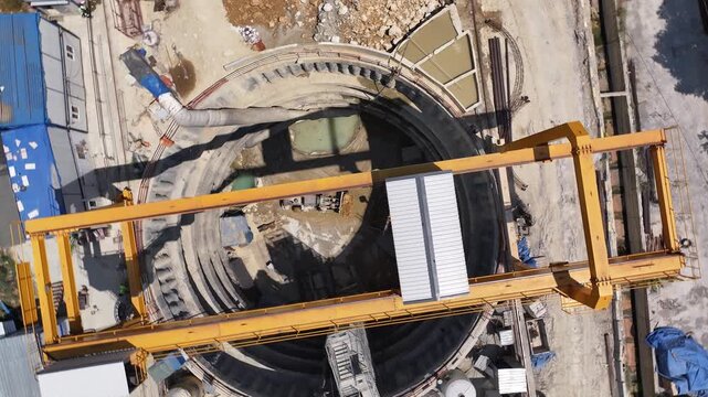 Top-down view of a metro construction shaft. Machinery working under the gantry crane shadow, circular bored pile wall, and site layout. Engineering and logistics operation detail.