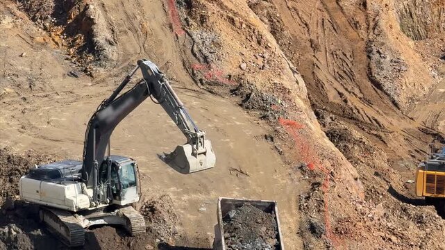 High-angle view of a white crawler excavator loading soil and rocks into a dump truck at a large site. Infrastructure preparation and urban development project.