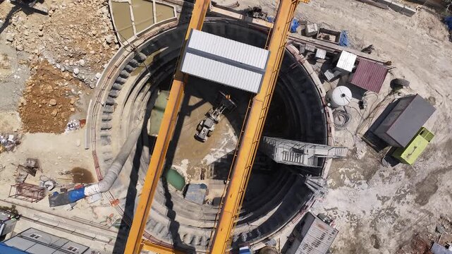 Top-down view of a metro construction shaft. Machinery working under the gantry crane shadow, circular bored pile wall, and site layout. Engineering and logistics operation detail.