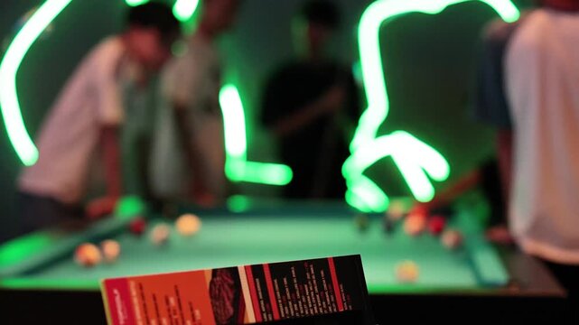 Blurry neon lights illuminate people playing pool in dimly lit room with menu in foreground.