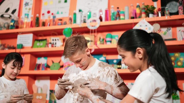 Caucasian student put clay on body while sculpt with carving equipment .Young happy girl smile while show hand with mud stick. Multicultural children playing with clay in pottery lesson. Edification