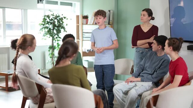 Teen group sit in circle and listen to Caucasian boy classmate speaking, teacher watching. Acting masters courses, stage speech courses, public speaking courses