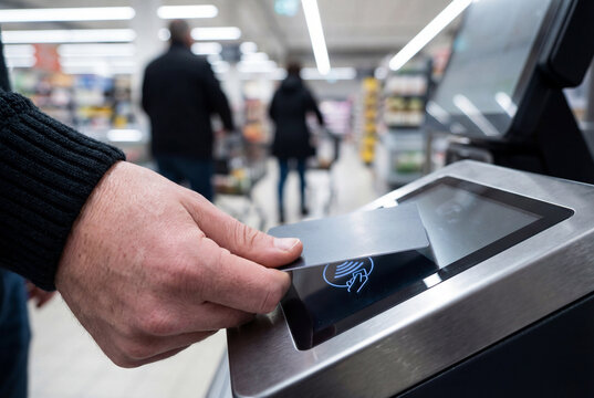 Close-up of a hand tapping a card on a self-checkout payment terminal in a supermarket. Cashless retail transaction concept with shallow depth of field