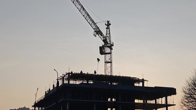Silhouette of a construction site with a towering crane and workers on a building frame against a sunset sky, with the sun peeking through the structure.