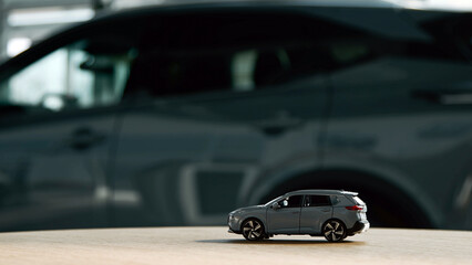 Fototapeta premium Miniature gray SUV model displayed on wooden surface with blurred full-sized gray SUV in the background, showcasing automotive design and scale comparison