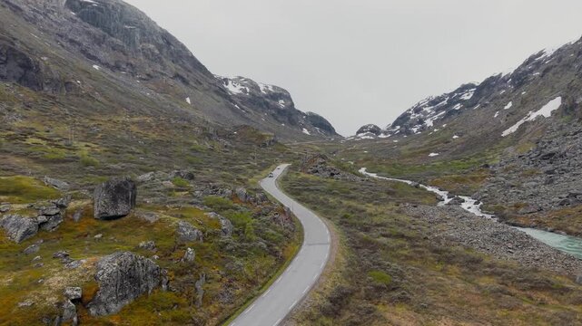 Drohnenflug &uuml;ber Stra&szlig;e neben Bach in Skandinavien, umgeben von verschneiten Berggipfeln, mosige Landschaft bei nebligem Wetter. RAW unbearbeitet